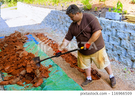 On construction site, worker digs trench for installation of drainage system containing perforated pipes. 116966509