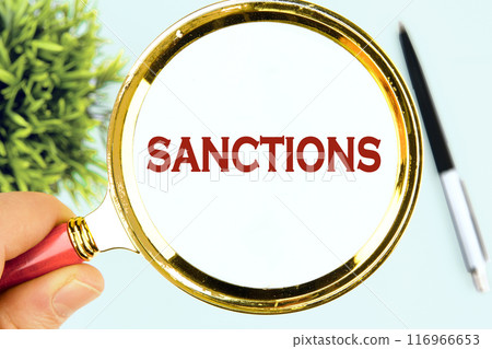SANCTIONS word written through a magnifying glass in his hand on a white surface SANCTIONS word written through a magnifying glass in his hand on a white surface 116966653