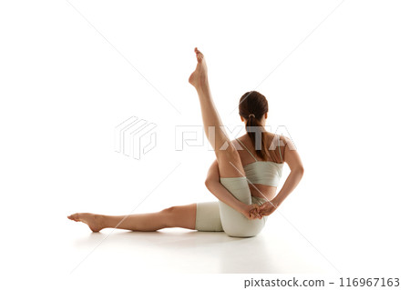 Fit woman practices seated leg stretch with twist against white studio background. Yoga training and overall physical fitness. 116967163
