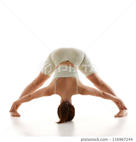 Young fit woman dressed comfy wearing, practicing wide-legged forward bend against white studio background, focuses on enhancing flexibility, relieving stress. 116967244