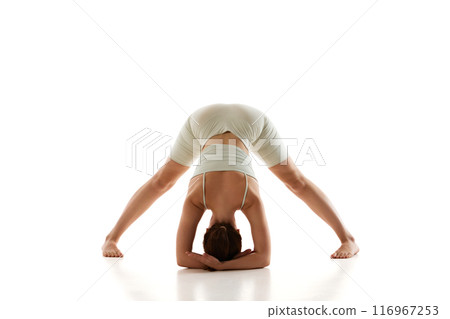 Young fit woman dressed comfy wearing, practicing wide-legged forward bend against white studio background, focuses on enhancing flexibility, relieving stress. 116967253