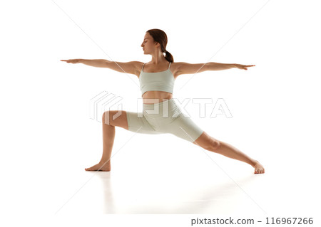 Woman in athletic wear performs yoga pose, with one knee bent and arms extended horizontally against white studio background. 116967266