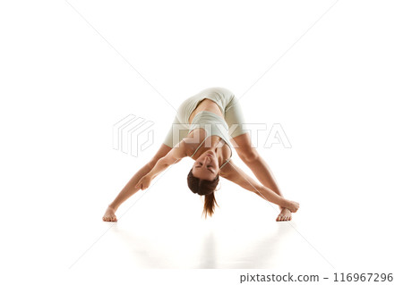 Woman in athletic wear performs wide-legged forward bend yoga pose, reaching one arm to side while keeping her head down against white studio background. 116967296
