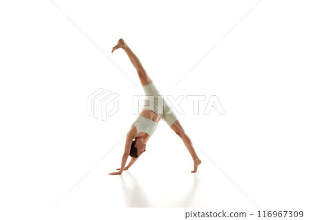 Sporty woman in athletic wear performs one-legged downward dog yoga pose demonstrating her flexibility against white studio background. 116967309
