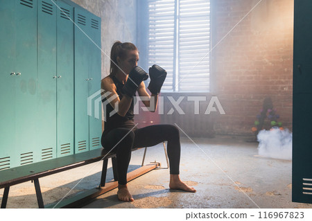 Workout in gym. Woman fighter ready for fight punching with boxing gloves in locker room. Strong girl training punches preparing for boxing sparring active exercise workout training in gym Workout in gym. Woman fighter ready for fight punching with boxing gloves in locker room. Strong girl training punches preparing for boxing sparring active exercise workout training in gym 116967823