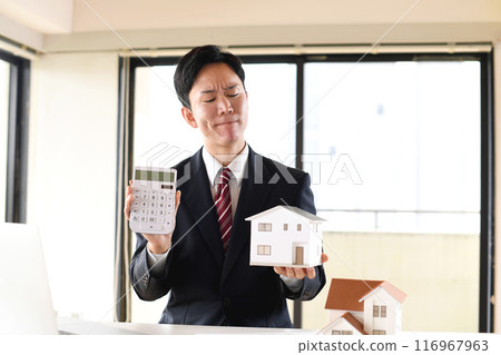 An Asian male business person in a suit is worried about a house model and a calculator 116967963