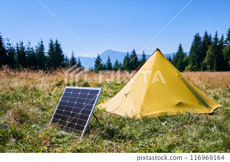 Yellow tent with solar panel set up in grassy field surrounded by dense evergreen trees under clear blue sky. Eco-friendly camping, integration of renewable energy in outdoor activities. Yellow tent with solar panel set up in grassy field surrounded by dense evergreen trees under clear blue sky. Eco-friendly camping, integration of renewable energy in outdoor activities. 116969164