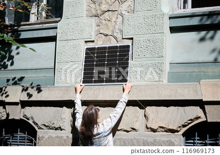 Woman holding photovoltaic solar panel in front of historical building, wearing plaid shirt and sunglasses. Concept of integration of sustainable renewable energy sources into old architecture. 116969173