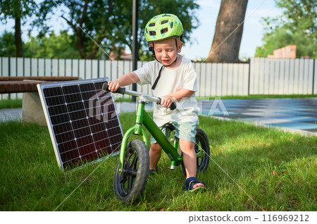 Young boy sits on green balance bike next to solar panel on grass. Kid wearing green helmet. Background features fence, trees, and bench. Concept of green energy generation. Young boy sits on green balance bike next to solar panel on grass. Kid wearing green helmet. Background features fence, trees, and bench. Concept of green energy generation. 116969212