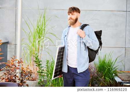 Bearded man carrying solar panel and black backpack, stands in front of a modern building. Happy guy in light blue shirt and white t-shirt surrounded by greenery and plants. 116969220