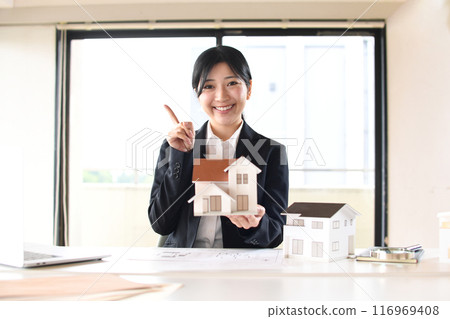 A female office worker holding a house model and shaking her index finger A female office worker holding a house model and shaking her index finger 116969408