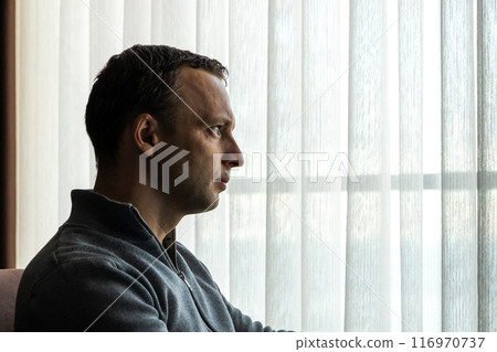 Serious young adult Caucasian man sitting in front of bright window 116970737