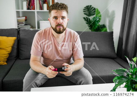 Young man spending time at home, sitting on a couch in apartment and playing arcade car video games on console. Male using controller to play street racing drift simulator. Young man spending time at home, sitting on a couch in apartment and playing arcade car video games on console. Male using controller to play street racing drift simulator. 116974274