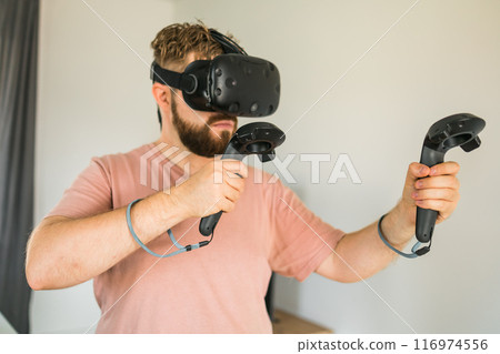Energetic young man wearing virtual reality headset and holding controllers plays in a video game at home. Active man playing VR quest adventure videogame of living room. 116974556
