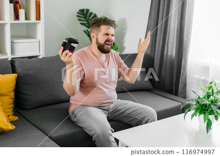 Cheerful young man seated on a sofa playing video games at home. Gaming addiction and entertainment 116974997