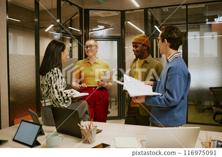 Group of four ethnically diverse young colleagues holding documents standing in modern office and having conversation 116975091