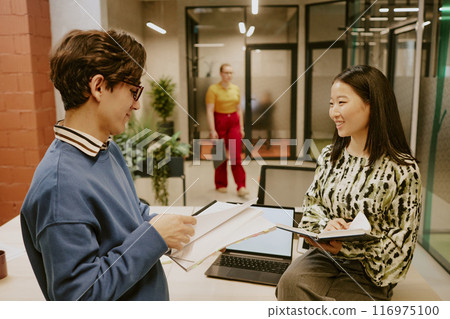 Young male and female managers wearing smart casual clothes holding notebooks in hands and discussing workflow 116975100
