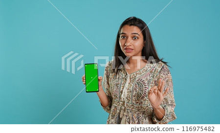 Woman presenting green screen mobile phone, isolated over studio background. Cheerful indian person holding copy space chroma key smartphone used for advertising brands, camera A Woman presenting green screen mobile phone, isolated over studio background. Cheerful indian person holding copy space chroma key smartphone used for advertising brands, camera A 116975482
