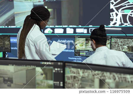 Manager offers logistics papers to indian employee in observation room, following delivery trucks on satellite navigation for routes around the city. Worker operating on CCTV public system. 116975509