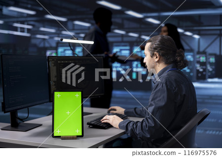Programmer works on green screen tablet in data center housing storage equipment. IT professional using mockup device in server farm with networking components providing vast amounts of computing Programmer works on green screen tablet in data center housing storage equipment. IT professional using mockup device in server farm with networking components providing vast amounts of computing 116975576