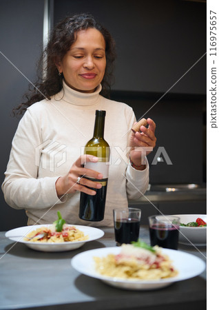 Woman opening a wine bottle while preparing dinner with pasta dishes in a cozy kitchen setting Woman opening a wine bottle while preparing dinner with pasta dishes in a cozy kitchen setting 116975657