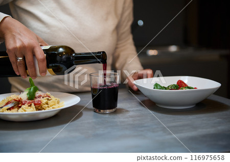 Person pouring red wine into a glass during a pasta dinner in a cozy home setting 116975658