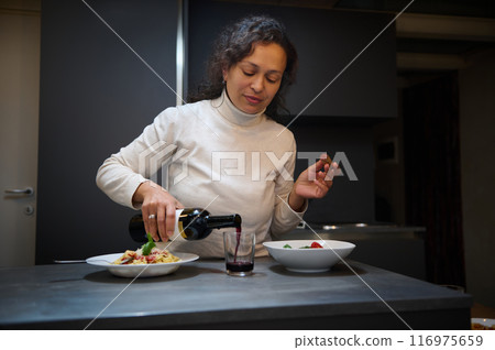 Woman pouring red wine into a glass while preparing a meal in a modern kitchen 116975659
