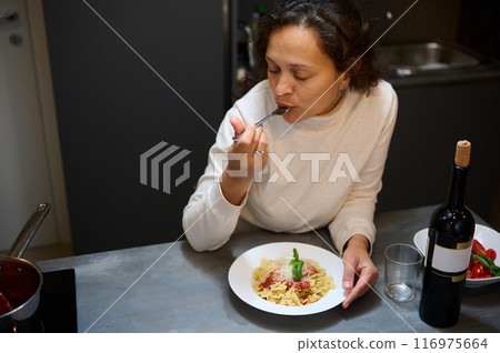 Woman enjoying a delicious pasta meal at home with a glass of wine in a cozy kitchen Woman enjoying a delicious pasta meal at home with a glass of wine in a cozy kitchen 116975664