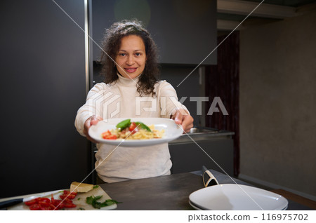 Smiling woman presenting a plate of pasta in a modern kitchen 116975702