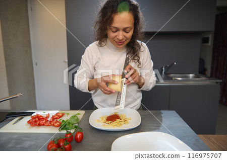 Woman grating cheese over pasta in a modern kitchen preparing an Italian meal Woman grating cheese over pasta in a modern kitchen preparing an Italian meal 116975707