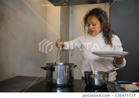 Woman cooking in a modern kitchen, stirring pots on the stove while holding plates 116975717