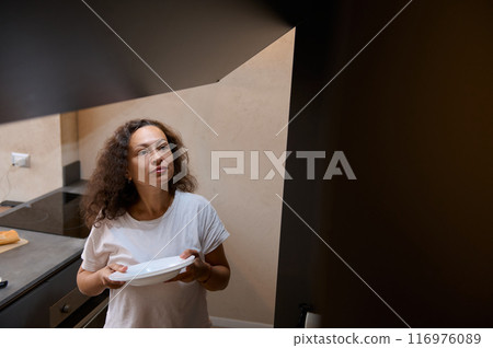 Woman in white t-shirt standing in kitchen holding a plate, looking thoughtful 116976089