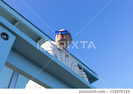 Smiling Middle Age Caucasian Male Standing Alone On Wooden Pier, Porch, Blue Sky on Background. Sunny Day. Man Wears Hooded Sweatshirt, Jeans, Sunglasses. Copy Space. Vacation Relaxation. Peaceful Smiling Middle Age Caucasian Male Standing Alone On Wooden Pier, Porch, Blue Sky on Background. Sunny Day. Man Wears Hooded Sweatshirt, Jeans, Sunglasses. Copy Space. Vacation Relaxation. Peaceful 116976148