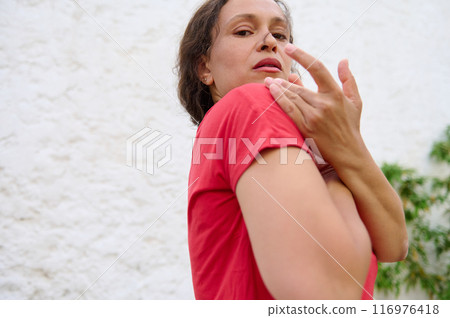 Woman in red shirt with thoughtful expression, posing outdoors against white wall background Woman in red shirt with thoughtful expression, posing outdoors against white wall background 116976418