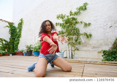 Woman practicing yoga outdoors on wooden deck with green plants in background Woman practicing yoga outdoors on wooden deck with green plants in background 116976456