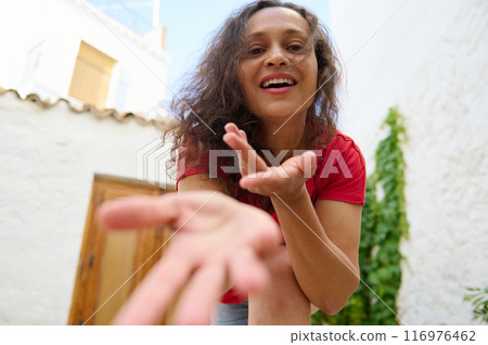 Cheerful young woman smiling and reaching out with open hands in bright outdoor setting 116976462