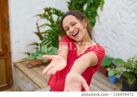 Happy woman in red shirt smiling joyfully and stretching hands towards camera outdoors 116976472