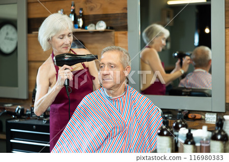 Female barber using hairdryer to style hair to elderly client in barbershop Female barber using hairdryer to style hair to elderly client in barbershop 116980383