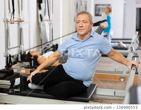 Senior male performing mermaid exercise on reformer Pilates machine in studio Senior male performing mermaid exercise on reformer Pilates machine in studio 116980553