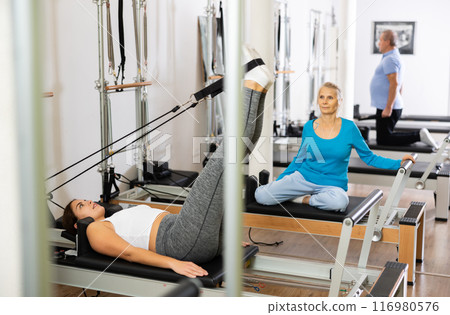 Positive young female concentrating on stretching exercise on Pilates chair equipment bed in studio. Active and healthy lifestyle concept 116980576