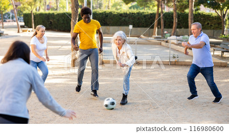 Joyful senior multinational friends playing soccer on a sandy playground on a warm spring evening Joyful senior multinational friends playing soccer on a sandy playground on a warm spring evening 116980600