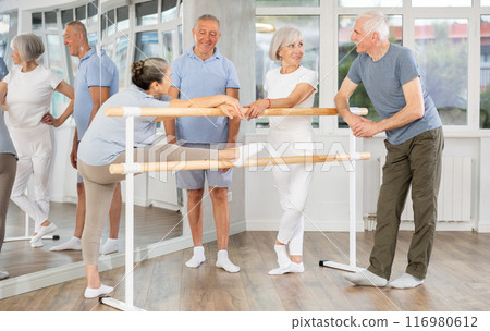 Warm-up sessions, mature women and men doing ballet exercise and talking, standing beside ballet barre in hall 116980612