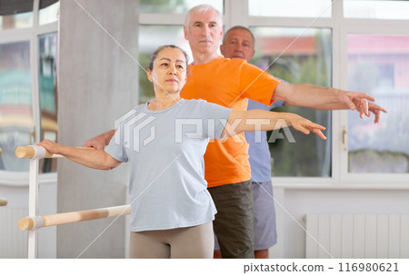 Pensioners of different ages exercise near ballet barre at group training session in two position Pensioners of different ages exercise near ballet barre at group training session in two position 116980621