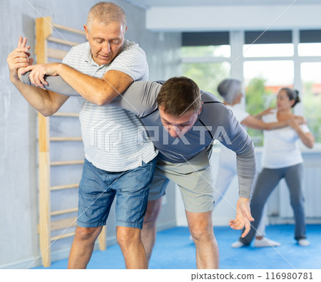 Senior man in pair with middle-aged coach train Krav Maga and self-defense skills. 116980781