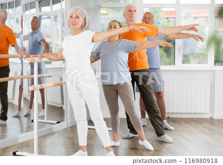 Group of positive aged women and men standing in row together while doing ballet dance moves, dancers standing in 2nd position of ballet stance in fitness room 116980918