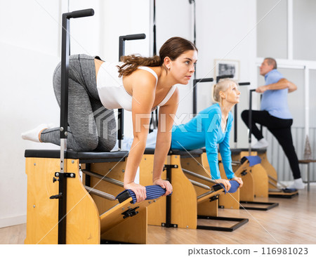 Young European woman working out on Pilates Wanda chair machine during group class with elderly people in gym Young European woman working out on Pilates Wanda chair machine during group class with elderly people in gym 116981023