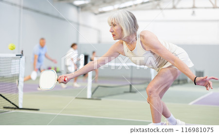 Elderly woman bouncing ball in pickleball game on court with racket Elderly woman bouncing ball in pickleball game on court with racket 116981075