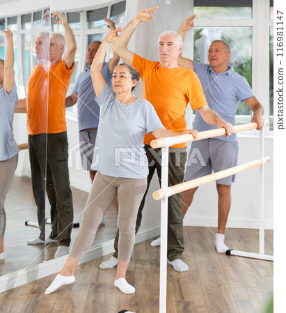 Active mature woman practicing battement tendu holding by ballet barre together with others 116981147