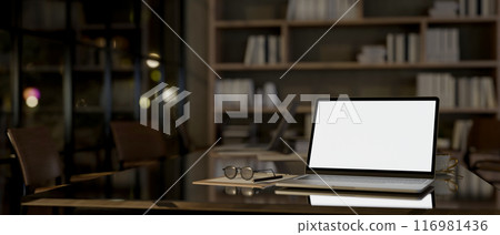 A laptop computer with a white-screen mockup on a meeting table in a modern meeting room at night. 116981436