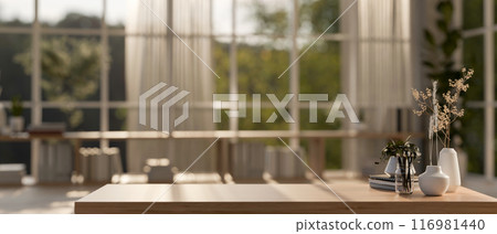 A wooden table in a contemporary Scandinavian room with sunlight streaming through a large window. 116981440